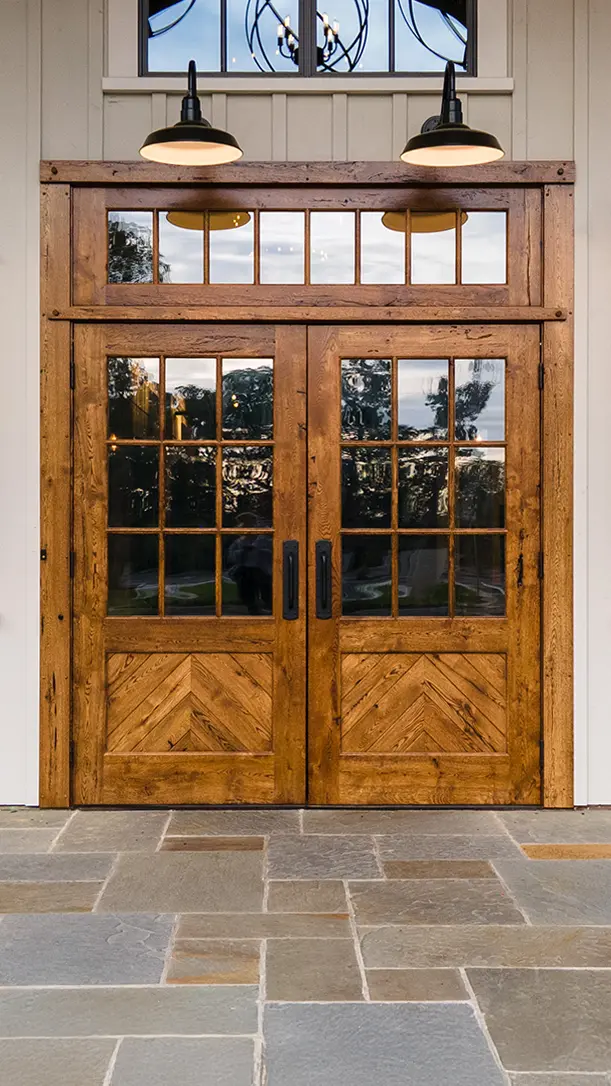 Double wooden doors with glass window panes and black handles, set in a white exterior wall. Two black outdoor lights are mounted above, and the doors open onto a stone tile patio. Trees are reflected in the glass.