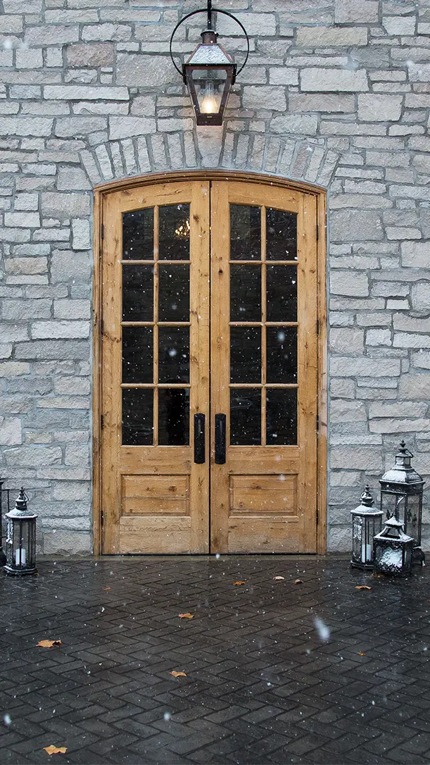 A pair of wooden double doors with glass panes set in a stone wall, flanked by metal lanterns. Snowflakes are falling, and a lantern hangs above the doorway. The ground is dark with a few leaves scattered.