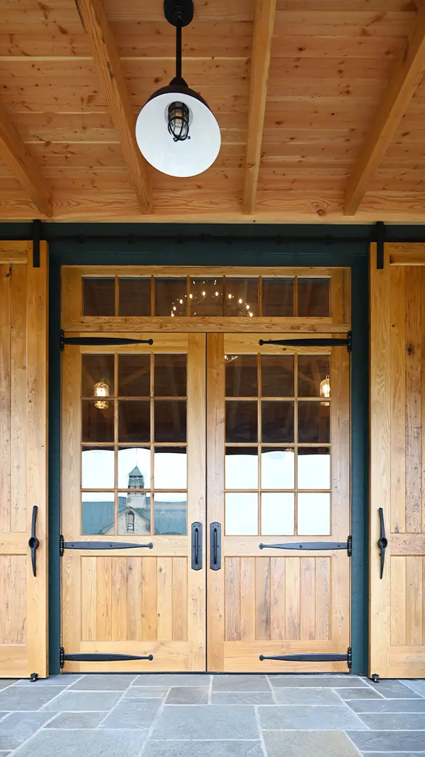 Double wooden doors with large glass panes and black metal hardware, set beneath a wooden porch ceiling with an industrial-style light fixture. A reflection of a building and sky is visible in the glass panes.