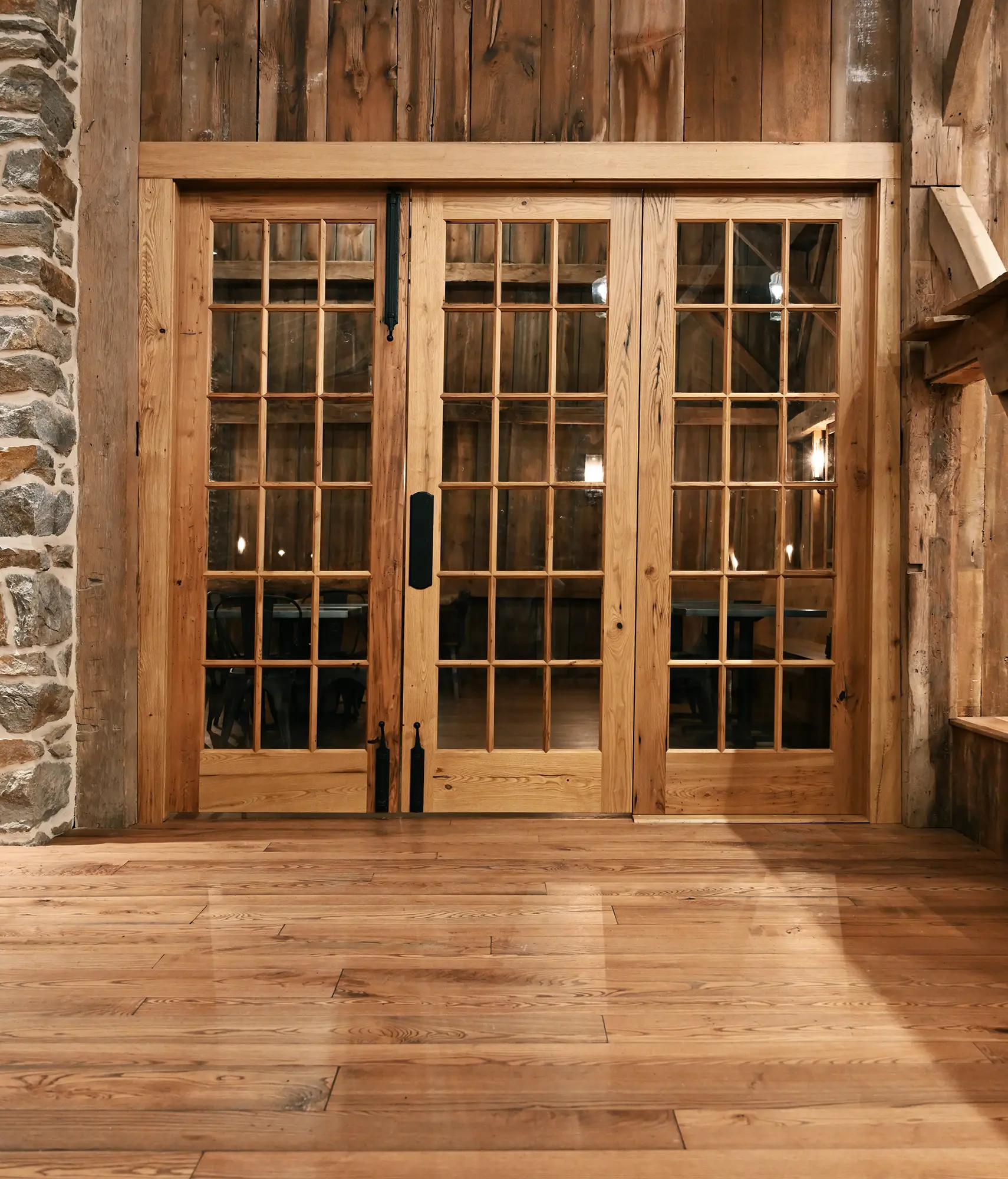 Wooden sliding barn doors with glass panes are set in a rustic interior, framed by a stone wall on the left and wooden walls and floor, with warm lighting visible through the glass.