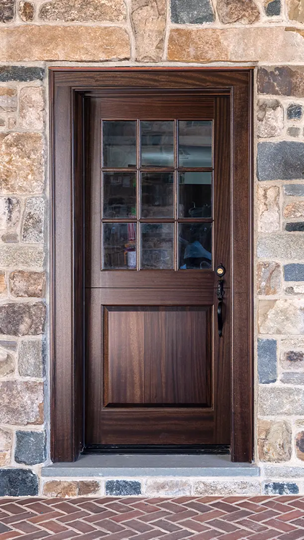 A wooden front door with a grid of six glass panes in the top half, set in a stone wall with various shades of brown and gray stones, above a herringbone brick doorstep.