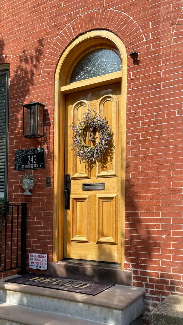 A wooden arched front door with a mail slot and a decorative wreath, set in a red brick building. The address 242 N Mulberry St is displayed beside the door. A small sign at the bottom requests package deliveries.