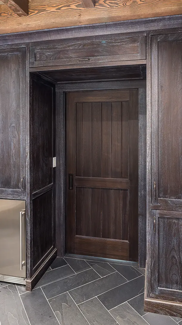 A dark wood paneled hidden door blends seamlessly with surrounding cabinetry in a modern kitchen, featuring a paneled design and matte black handle. The floor is covered in dark herringbone tiles.