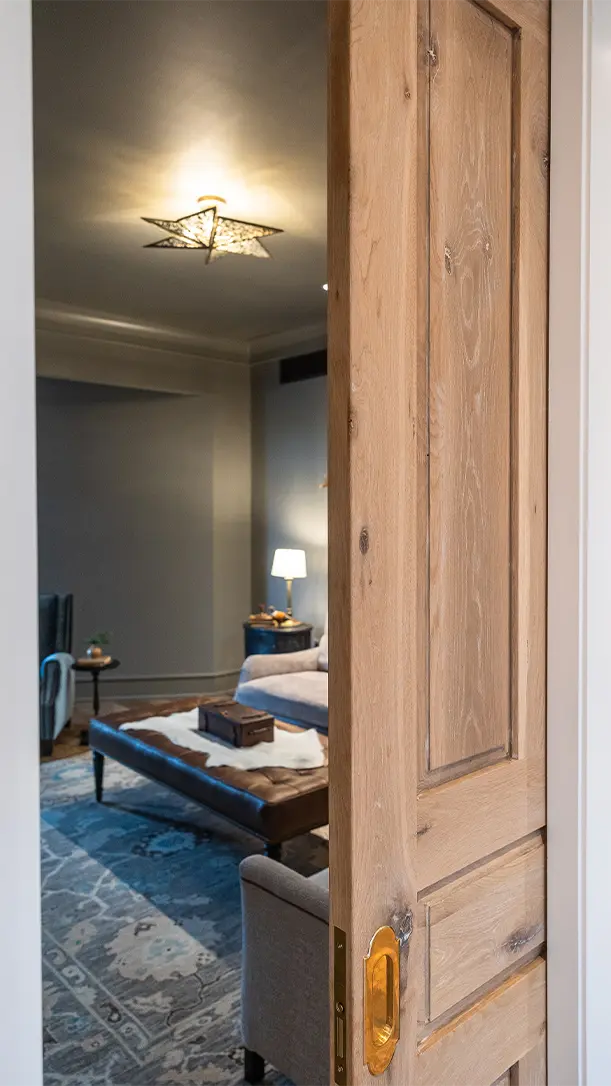 A partially open wooden door reveals a cozy living room with a patterned rug, a sofa, a coffee table, and a lit table lamp in a softly lit, neutral-toned space. A star-shaped ceiling light is visible above.