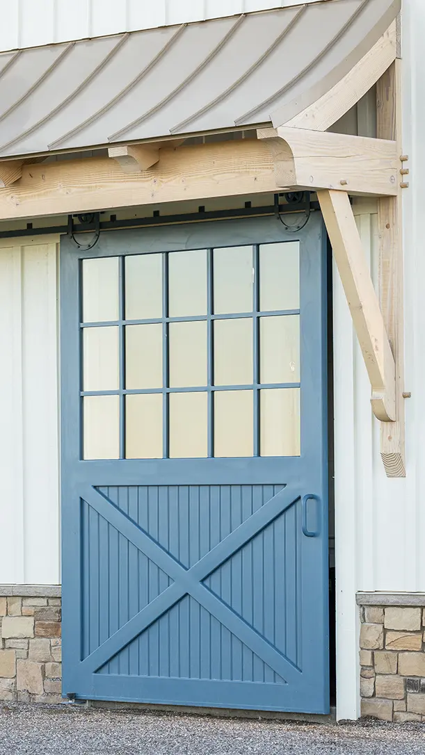 A partially open blue sliding barn door with glass window panes, set in a white building with stone trim and exposed wooden supports under a metal roof.