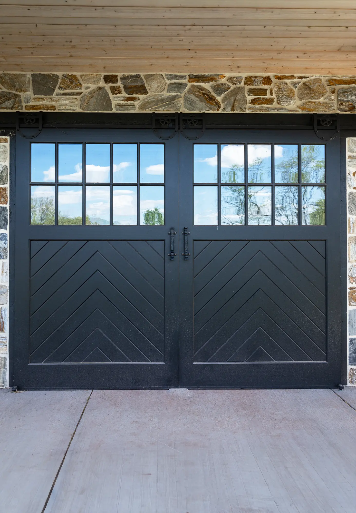 Black double barn-style garage doors with diagonal wood patterns and window panes at the top, set in a stone wall under a wood-paneled ceiling. Reflections of trees and sky are visible in the glass.