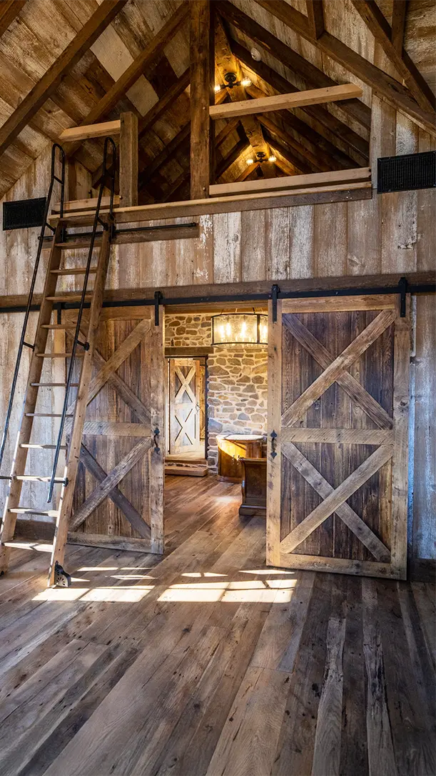 Rustic interior with wooden floors, sliding barn doors, exposed beams, a wooden ladder leading to a loft, and stone walls visible in the next room lit by a chandelier.