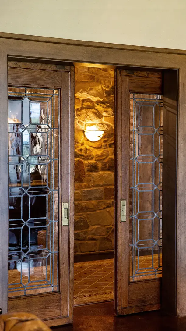 Wood-framed glass sliding doors with geometric patterns are partially open, revealing a warmly lit room with stone walls and a mounted wall sconce in the background.