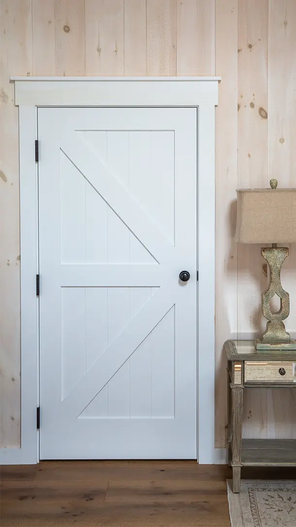 A white wooden door with a diagonal cross design is set in a light wood-paneled wall. To the right, there is a rustic side table with a beige lamp on top. The floor is medium-toned wood.