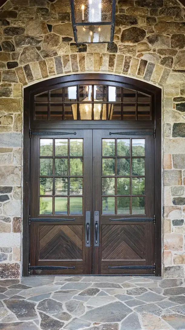 Large double wooden front doors with glass panes, framed by a stone wall and topped with transom windows. A hanging lantern above the doors adds a classic touch. Reflections of trees can be seen in the glass.