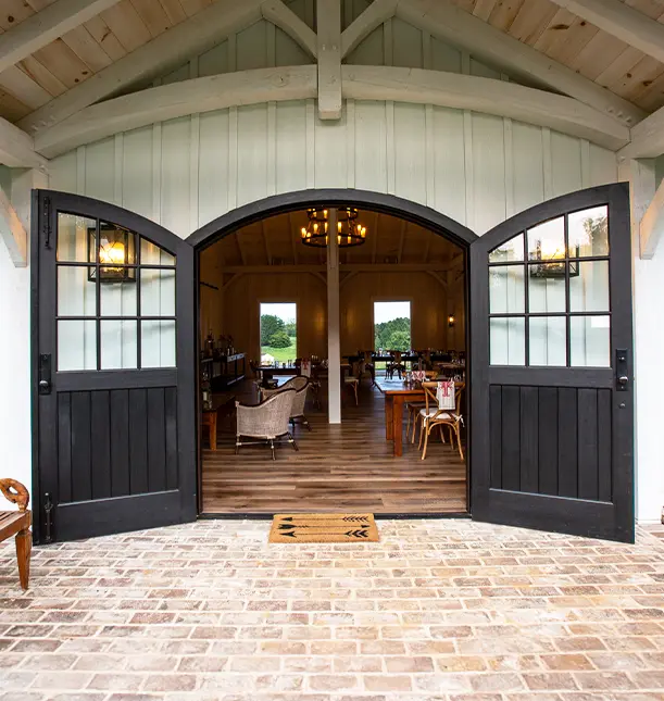 Open double barn doors with black frames lead into a warmly lit rustic venue with wooden floors, chandeliers, and seating areas. The entryway is brick, and greenery is visible through windows at the back.