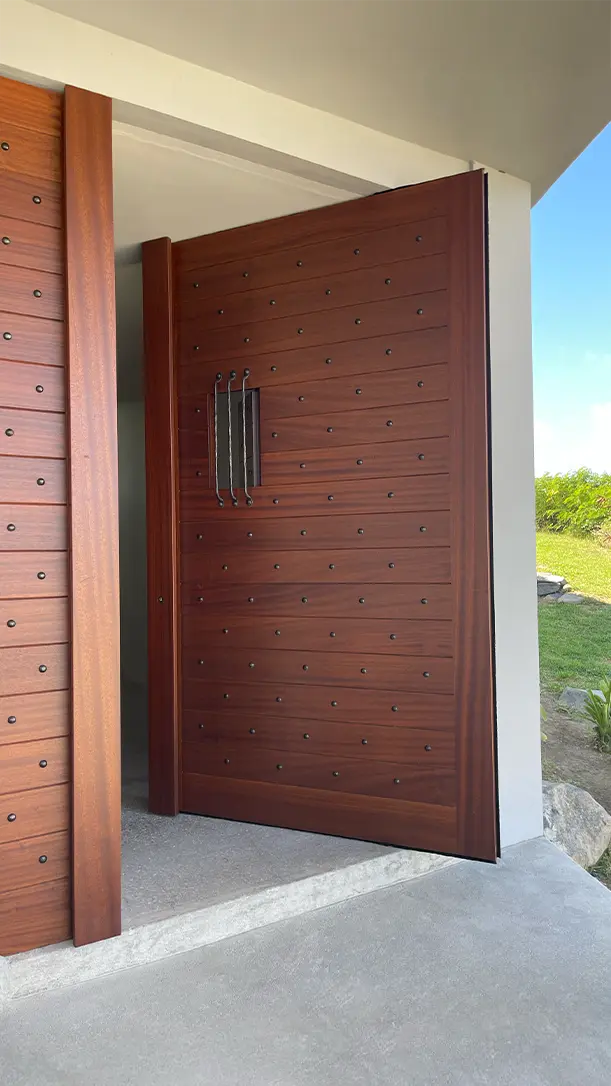A large, modern wooden front door with metal studs and a vertical handle is partially open, revealing a green lawn and blue sky outside. The door is set in a contemporary entryway with concrete flooring.