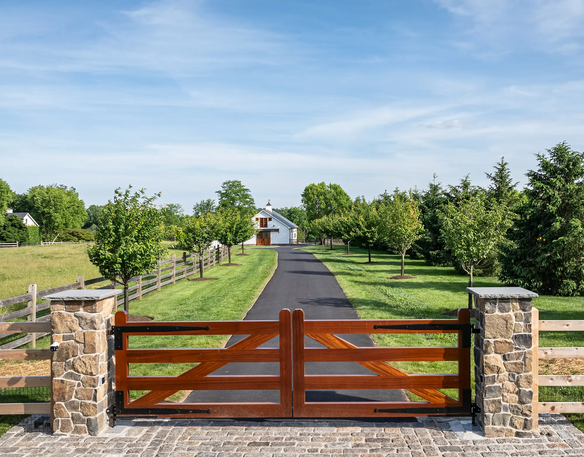 A driveway with a wooden gate and stone pillars leads through green lawns and trees to a house in the distance under a blue sky.