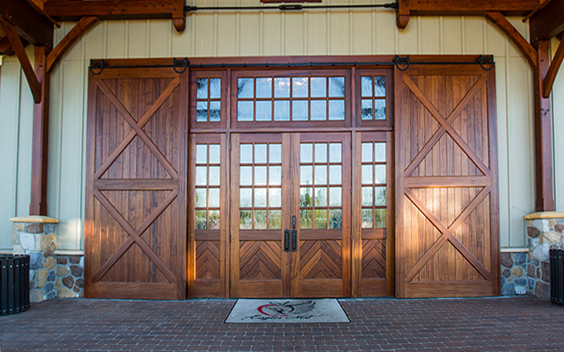 Large wooden barn doors with diagonal planks flank a set of double glass doors and windows. The entrance sits on brick pavement with a welcome mat in front, supported by stone pillars and wood beams.