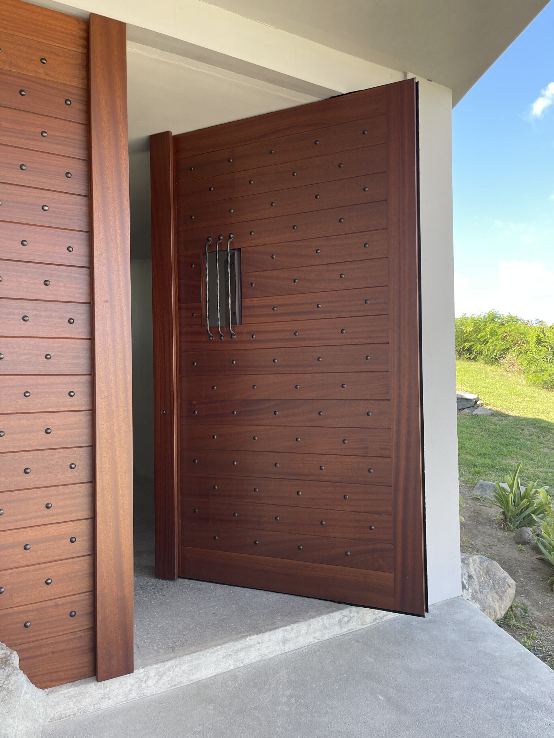 A large, modern wooden front door with metal studs and a vertical handle is partially open, revealing a green lawn and blue sky outside. The door is set in a contemporary entryway with concrete flooring.
